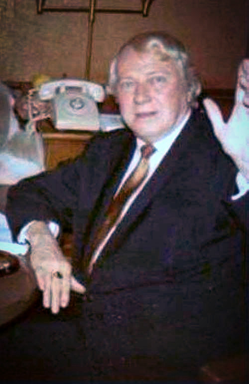 Criswell seated in office with telephone, and waving.